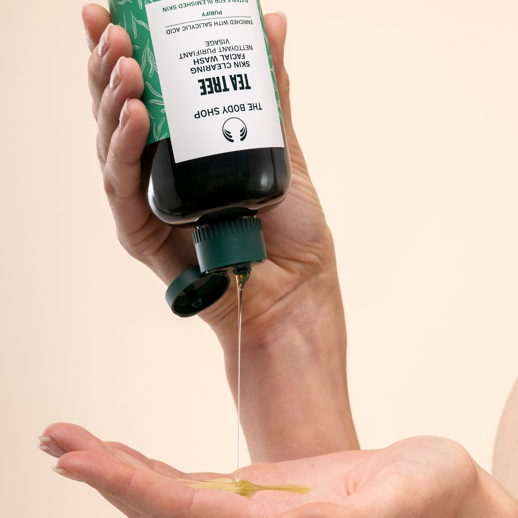 Hand pouring The Body Shop Tea Tree Facial Wash from a bottle into another hand against a beige background.